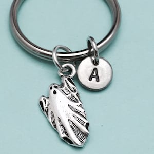 May include: A silver-colored keychain with a ghost charm and a circular charm with the letter "A". The ghost charm has etched details. The keychain is on a light blue background.