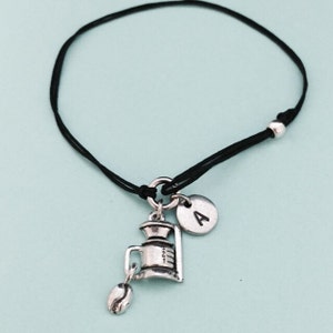 Puede incluir: Un brazalete de cordón negro con un dije de plata de un molinillo de café y un grano de café. El dije también tiene un disco de plata con la letra 'A' en él.