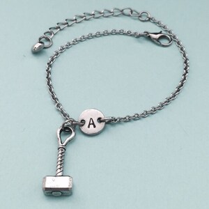 May include: A silver chain bracelet with a silver charm of a hammer and a silver disc with the letter "A" on it.