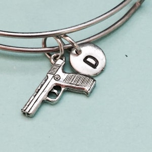 Op de afbeelding: Zilveren armband met een pistoolbedel en een letter D-bedel. Het armband is op een lichtblauwe achtergrond.