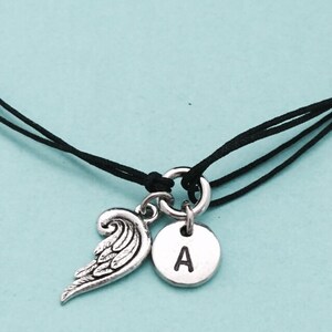 May include: A black cord bracelet with a silver charm featuring an angel wing and a round disc with the letter 'A' engraved on it.