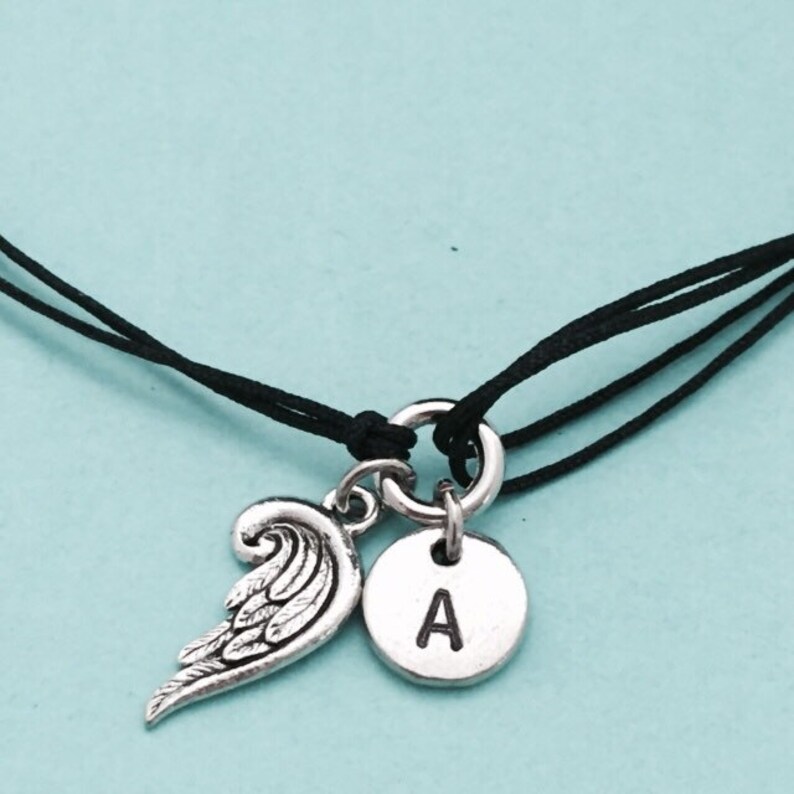 May include: A black cord bracelet with a silver charm featuring an angel wing and a round disc with the letter 'A' engraved on it.