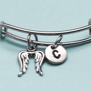 May include: A silver bangle bracelet with two charms. One charm is a pair of angel wings and the other is a round charm with the letter "C" on it.