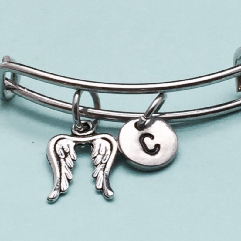 May include: A silver bangle bracelet with two charms. One charm is a pair of angel wings and the other is a round charm with the letter "C" on it.