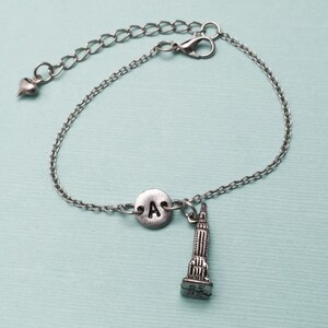 May include: A silver chain bracelet with a charm of the Empire State Building and a silver disc with the letter 'A' on it.
