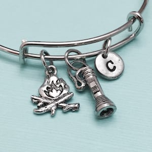 May include: Silver-toned camping-themed charm bracelet. The bracelet features charms of a campfire, a flashlight, and a circular charm with the letter "C". The bracelet is on a light blue background.