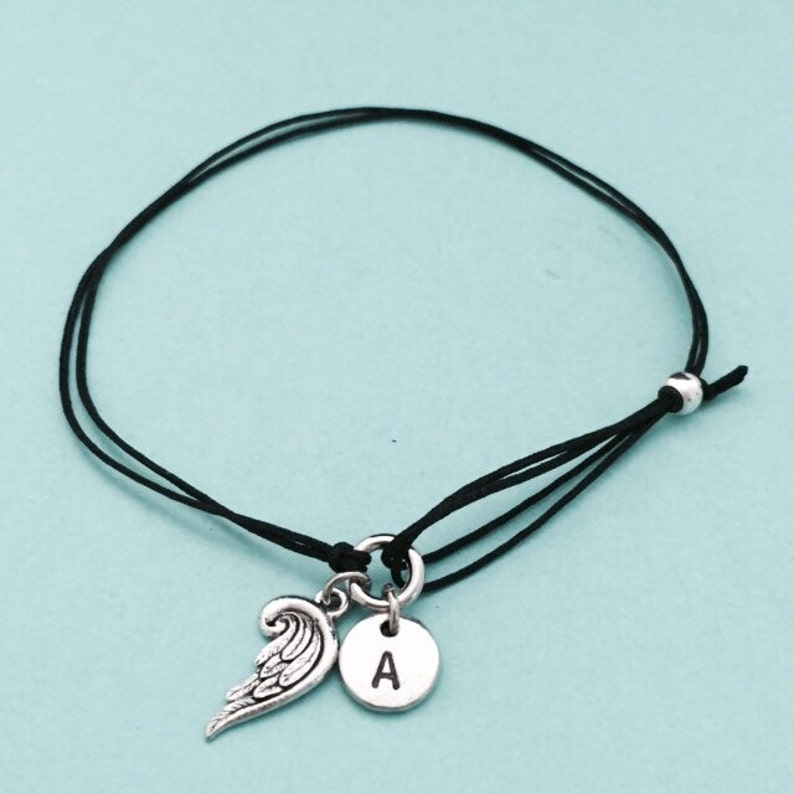May include: A black string bracelet with a silver angel wing charm and a silver charm with the letter A.