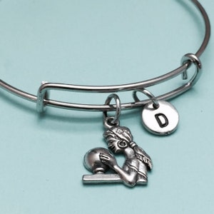 May include: Silver-toned bangle bracelet with a charm featuring a woman holding a sphere. A small, round charm with the letter "D" is also attached. The bracelet is set against a light blue background.