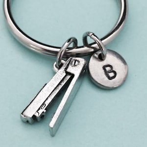 Op de afbeelding: Een zilveren sleutelhanger met een miniatuur nietmachine-bedel, een ronde bedel met de letter "B" erop gegraveerd en een zilveren sleutelring.