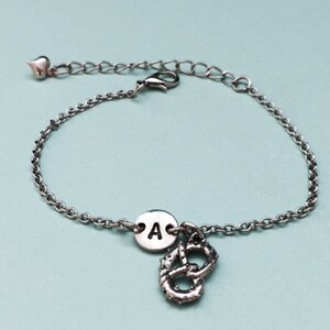 May include: A silver chain bracelet with a charm of a pretzel and a round charm with the letter A.