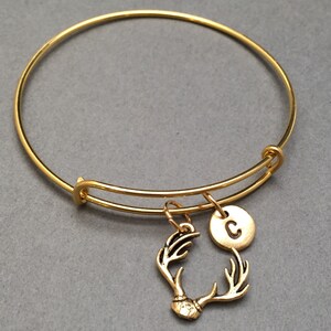 May include: A gold bangle bracelet with a charm of antlers and a round charm with the letter "C" engraved on it.