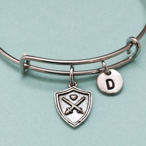 Op de afbeelding: Een zilveren armband met een schildbedel met gekruiste zwaarden en een hart, en een bedel met de letter 'D'. De armband is op een lichtblauwe achtergrond.