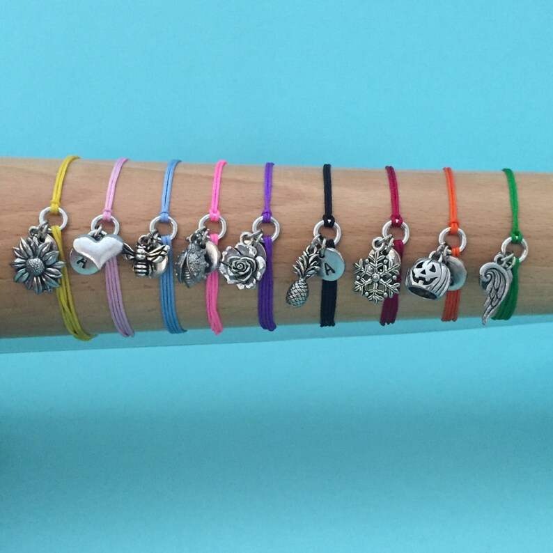 May include: Ten colorful friendship bracelets with different charms, including a sunflower, heart, bee, owl, rose, pineapple, letter A, snowflake, pumpkin, and angel wing.