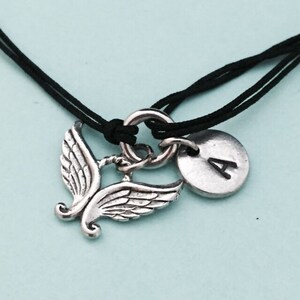 May include: A black cord bracelet with a silver charm featuring a pair of angel wings and a round disc with the letter 'A' engraved on it.