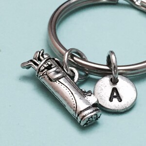 May include: Silver golf bag keychain with a silver initial charm. The initial charm has the letter "A" engraved on it.
