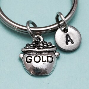 May include: Silver-toned keychain featuring a pot of gold charm with the word "GOLD" and a round charm with the letter "A". The charms are attached to a silver ring. The background is a light blue color.