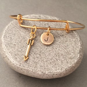 Op de afbeelding: Gouden armband met een hanger in de vorm van een pijl en een hanger met de letter J.