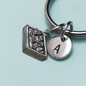 May include: Silver keychain with a small book charm and a round charm with the letter A. The book charm has a detailed design of a person reading.
