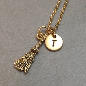 Puede incluir: Un collar dorado con un pequeño dije de escoba y un dije redondo con la letra "T". El dije de la escoba tiene detalles intrincados, y la cadena del collar es delicada. El colgante está suspendido de una cadena dorada, creando un accesorio caprichoso.
