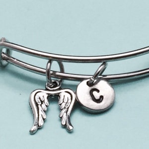 May include: A silver bangle bracelet with two charms: a pair of angel wings and a round charm with the letter "C" engraved on it.