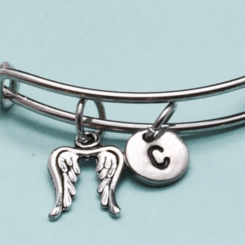 May include: A silver bangle bracelet with two charms: a pair of angel wings and a round charm with the letter "C" engraved on it.