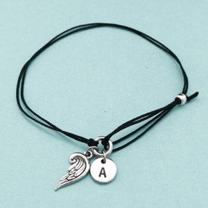 May include: A black string bracelet with a silver charm featuring an angel wing and a letter 'A'.
