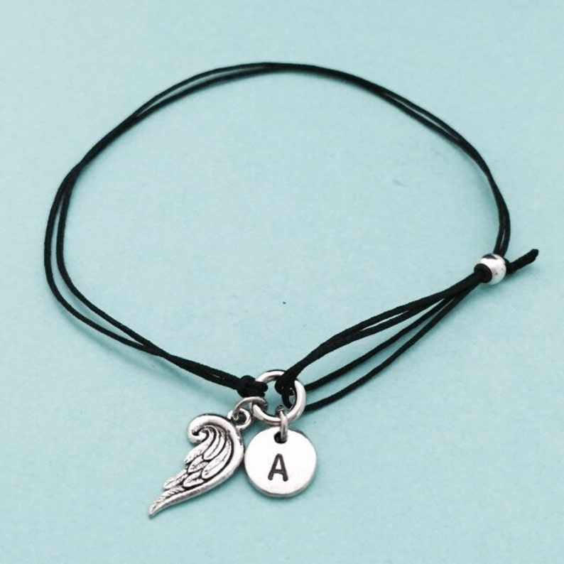 May include: A black string bracelet with a silver charm featuring an angel wing and a letter 'A'.