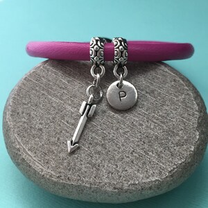 Op de afbeelding: Een roze leren armband met twee zilveren bedels. Een bedel is een kleine pijl en de andere is een ronde bedel met de letter "P" erop gegraveerd.