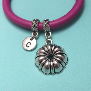 May include: A close-up of a purple bracelet with two silver charms. One charm is a small, round disc with the letter "C" on it. The other charm is a silver flower shape with a dark green center.