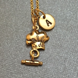 May include: Gold necklace with a chef hat charm, a rolling pin charm, and a letter 'A' charm. The chef hat charm has the words 'le chef' inscribed on it.