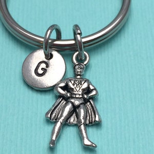 May include: Silver-coloured keyring featuring a superhero charm with a cape, standing with hands on hips. Includes a round charm with the letter "G". The keyring is on a silver ring, set against a light blue background.