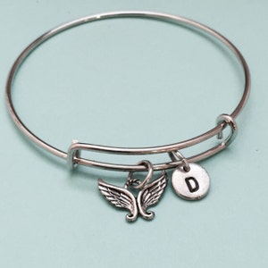 May include: A silver bangle bracelet with a charm featuring a pair of angel wings and a silver disc with the letter "D" engraved on it.