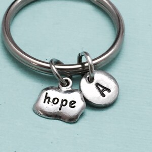May include: Silver-toned keychain with a large ring. It features two charms: one shaped like a pebble with the word "hope" and a round charm with the letter "A". The charms hang from the ring.