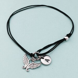 May include: A black cord bracelet with a silver angel wing charm and a silver disc charm with the letter A.