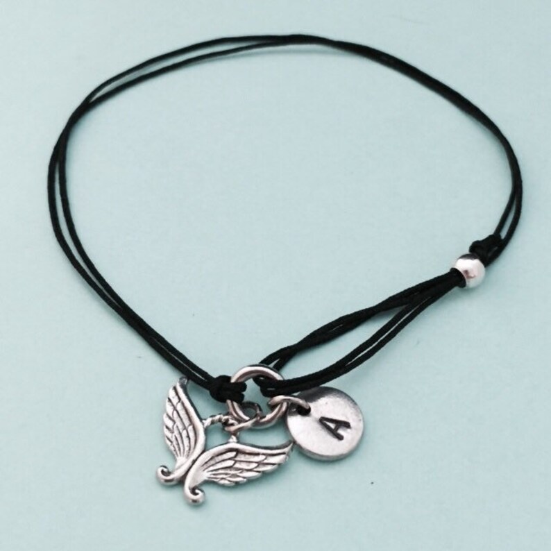 May include: A black cord bracelet with a silver angel wing charm and a silver disc charm with the letter A.