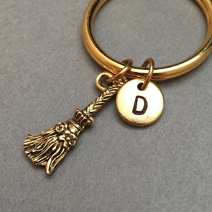 Puede incluir: Llavero dorado con un pequeño amuleto de escoba y un amuleto redondo con la letra "D" grabada.