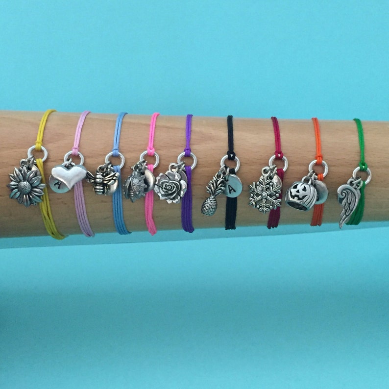 May include: Ten colorful string bracelets with different silver charms, including a sunflower, heart, bee, rose, pineapple, letter A, snowflake, pumpkin, and angel wing.
