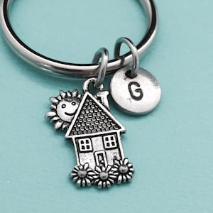 Puede incluir: Llavero de plata con un dije de casa, un sol sonriente y un dije de la letra G. El dije de la casa tiene una pequeña puerta y ventanas.