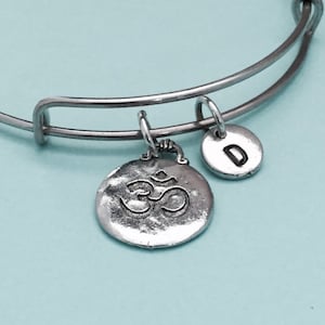 Puede incluir: Pulsera de brazalete plateada expandible con dos colgantes. Uno es un colgante redondo con el símbolo Om, y el otro es un pequeño disco con la letra "D". La pulsera está sobre un fondo azul claro.