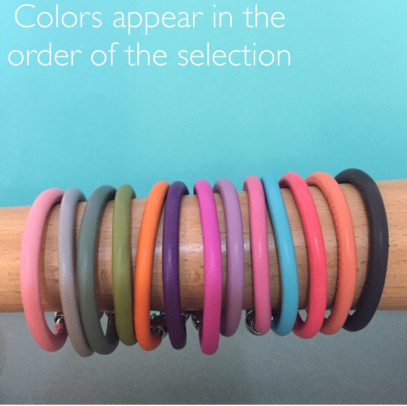 May include: A set of ten leather bracelets in various colors, including gray, green, orange, purple, pink, lavender, turquoise, coral, brown, and black. The bracelets are arranged in a row on a wooden surface. The text "Colors appear in the order of the selection" is visible above the bracelets.