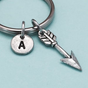 May include: Silver-toned keychain with a round charm featuring the letter "A" and an arrow charm. The arrow has a detailed feather design. The keychain is on a silver ring, perfect for keys or bags.