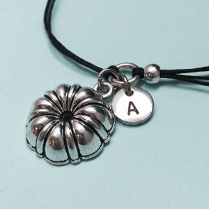 May include: A silver charm necklace with a small cake pan charm and a round charm with the letter "A" engraved on it. The necklace is on a black cord.