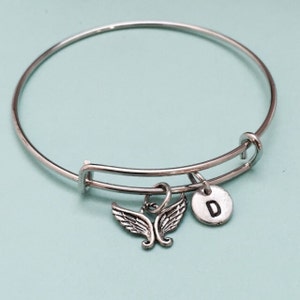 May include: A silver bangle bracelet with a charm featuring a pair of angel wings and a small round charm with the letter "D" engraved on it.