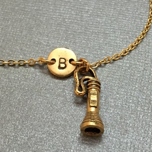 May include: Gold necklace with a charm of a flashlight and a round charm with the letter B.