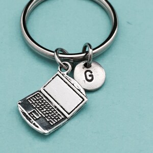 Könnte beinhalten: Silberner Laptop-Schlüsselanhänger mit einem silbernen Initial-Anhänger. Der Initial-Anhänger ist der Buchstabe "G".