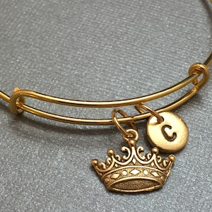 May include: Gold expandable bangle bracelet with a gold crown charm and a gold charm with the letter C.