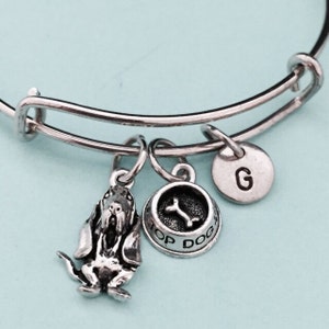 Puede incluir: Pulsera extensible de plata con abalorios. Los abalorios incluyen un perro, un cuenco para perros con un hueso y una letra G.