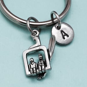May include: Silver-toned keychain featuring a ski lift charm with two figures, a ski, and a round charm with the letter "A". The keychain is attached to a silver ring. The ski lift charm has intricate details.