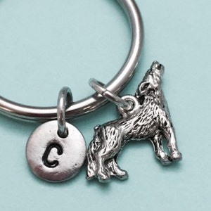 May include: A silver-colored keychain featuring a howling wolf charm and a small round charm with the letter "C". The keychain has a silver ring for attaching keys. The wolf charm is detailed with textured fur.