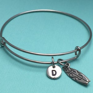 Op de afbeelding: Een zilveren, verstelbare armband met een bedel in de vorm van een surfboard en een bedel met de letter "D" erop.